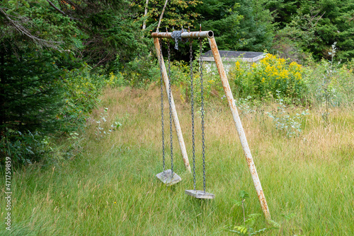 An old rusty homemade children's swing set. The playground equipment is made of white rusty metal pipes, chains, and wooden seats. There are two swings hanging from the play equipment in a field.