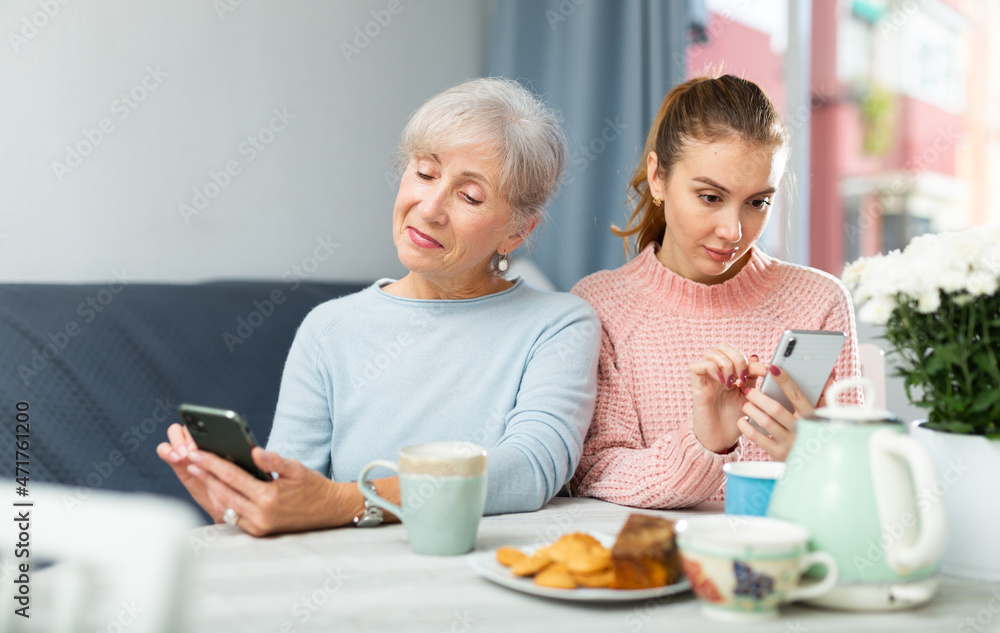Positive senior woman and her adult daughter sitting at table in cozy dining room, looking absorbedly at cell phones . Concept of family phubbing