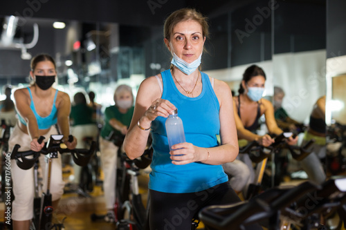 Mature female in fase mask drinking water during cycling class in modern gym