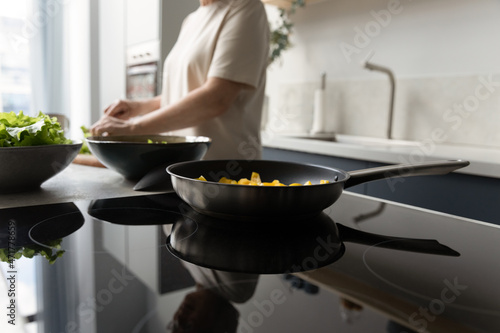 Frying pan with vegetable meal on induction cooker close up. Woman cooking dinner, preparing salad, slicing fresh vegetables into bowl with lettuce. Culinary, kitchen utensil, appliance concept photo