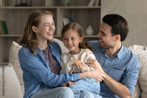 Fotografie Overjoyed young couple parents tickling adorable little kid daughter, enjoying entertaining domestic carefree playtime activity, resting together on comfortable sofa at home, loving relations concept