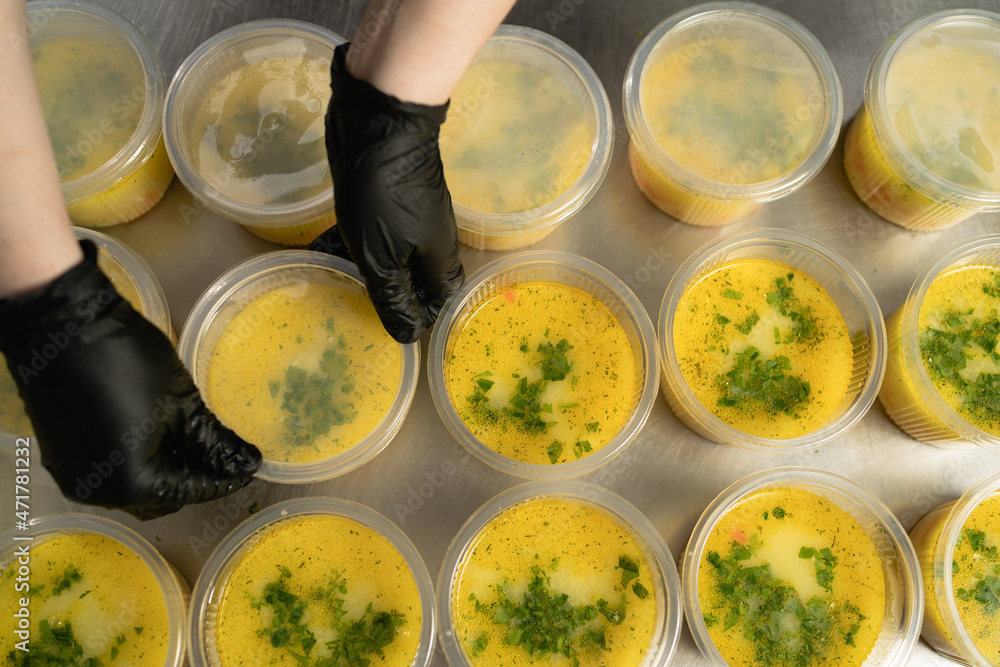 many disposable plates with soup in a lunch box stand on the table in the kitchen in a restaurant female hands are covering the food with a lid. Takeaway food concept during quarantine or pandemic. many disposable plates with soup in a lunch box stand on the table in the kitchen in a restaurant female hands are covering the food with a lid. Takeaway food concept during quarantine or pandemic.