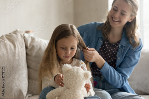 Happy cute small kid girl playing with favorite toy while caring affectionate mother combing her blonde light hair, getting ready for kindergarten or school, doing hairstyle, sitting on couch at home.