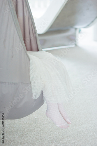 Close up girls legs in white tights and white dress. Cropped view of little girl relaxing in hanging grey chair bag or hammock in light room with carpet. Swing at home.