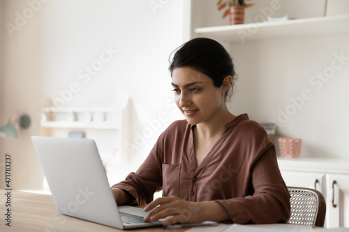 Smiling young indian ethnicity woman working on computer, sitting at table in modern light home office, creating new project, typing communicating distantly with client, web surfing information online