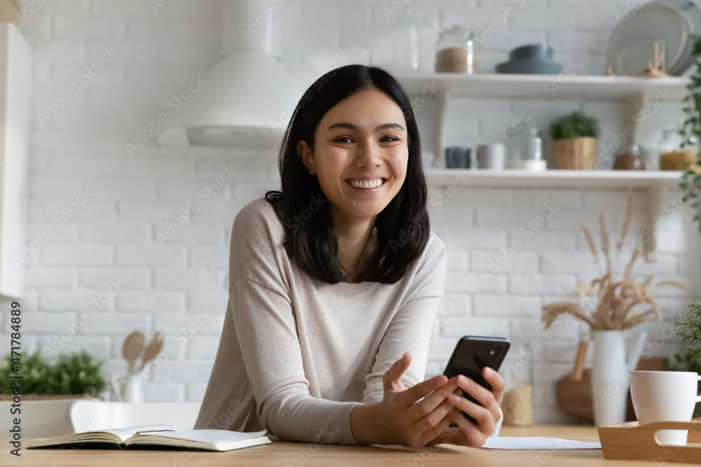 Attractive young Asian woman sit in kitchen holds smartphone smile look at camera, pose with wireless device in hands. Happy client of mobile app food delivery services, ecommerce, modern tech concept Attractive young Asian woman sit in kitchen holds smartphone smile look at camera, pose with wireless device in hands. Happy client of mobile app food delivery services, ecommerce, modern tech concept