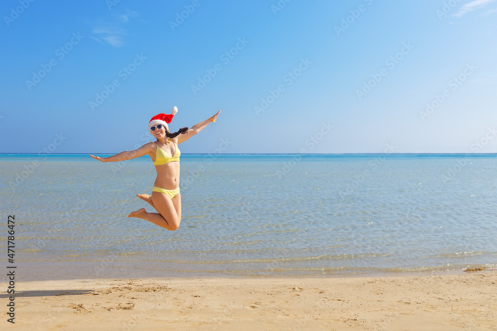 happy woman in santa hat and sunglasses jumping on sand beach