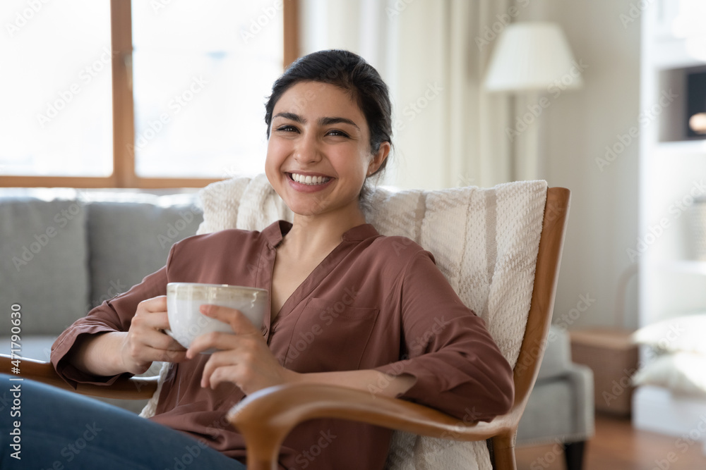 Portrait of smiling beautiful indian ethnicity woman resting in cozy armchair with cup of coffee or tea in hands, enjoying carefree lazy relaxed morning weekend time lone in modern living room. Portrait of smiling beautiful indian ethnicity woman resting in cozy armchair with cup of coffee or tea in hands, enjoying carefree lazy relaxed morning weekend time lone in modern living room.