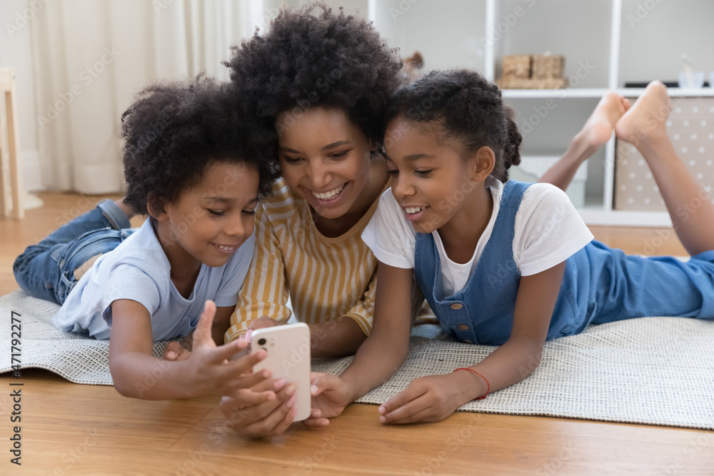 African American nanny teaching kids to use online app on smartphone. Happy young mother and sibling children lying on warm floor, making video call on mobile phone, watching content, smiling African American nanny teaching kids to use online app on smartphone. Happy young mother and sibling children lying on warm floor, making video call on mobile phone, watching content, smiling