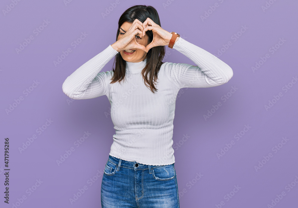 Beautiful brunette woman wearing casual clothes doing heart shape with hand and fingers smiling looking through sign Beautiful brunette woman wearing casual clothes doing heart shape with hand and fingers smiling looking through sign