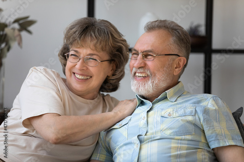 Happy senior married couple home candid portrait. Cheerful middle aged husband and wife resting on couch at home, hugging, looking away, smiling, laughing, enjoying leisure time together