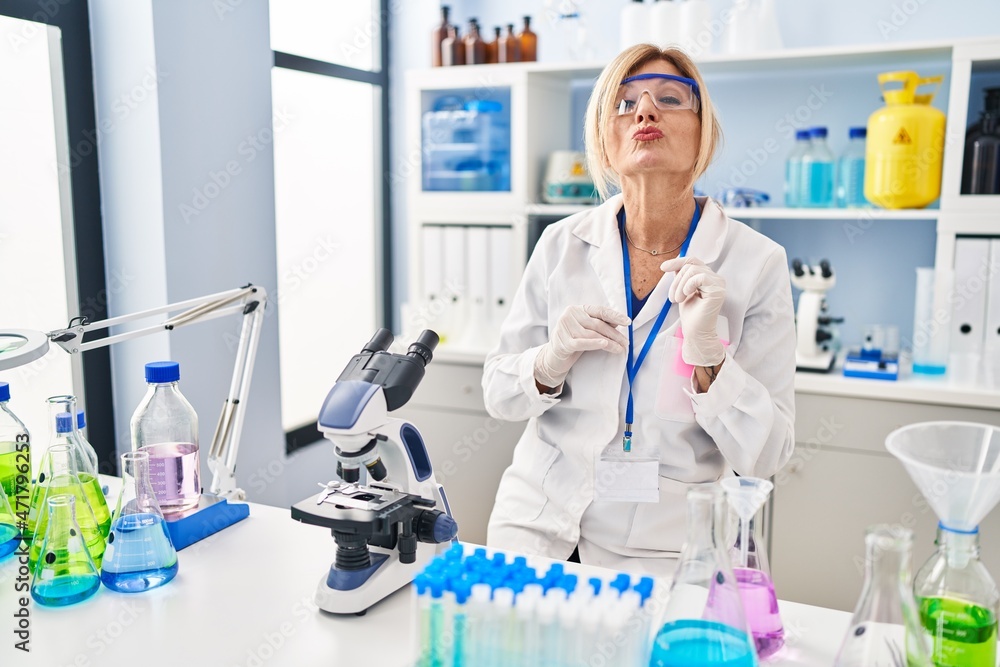 Middle age blonde woman working at scientist laboratory looking at the camera blowing a kiss on air being lovely and sexy. love expression. Middle age blonde woman working at scientist laboratory looking at the camera blowing a kiss on air being lovely and sexy. love expression.