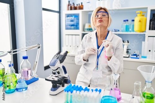 Middle age blonde woman working at scientist laboratory looking at the camera blowing a kiss on air being lovely and sexy. love expression.