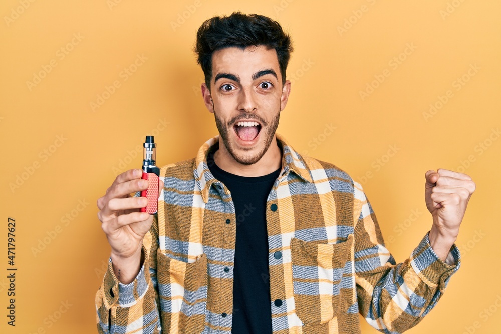 Young hispanic man holding electronic cigarette screaming proud, celebrating victory and success very excited with raised arms Young hispanic man holding electronic cigarette screaming proud, celebrating victory and success very excited with raised arms