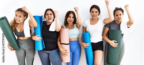 Group of women holding yoga mat standing over isolated background angry and mad raising fist frustrated and furious while shouting with anger. rage and aggressive concept.
