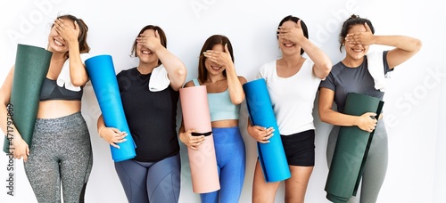 Group of women holding yoga mat standing over isolated background smiling and laughing with hand on face covering eyes for surprise. blind concept.