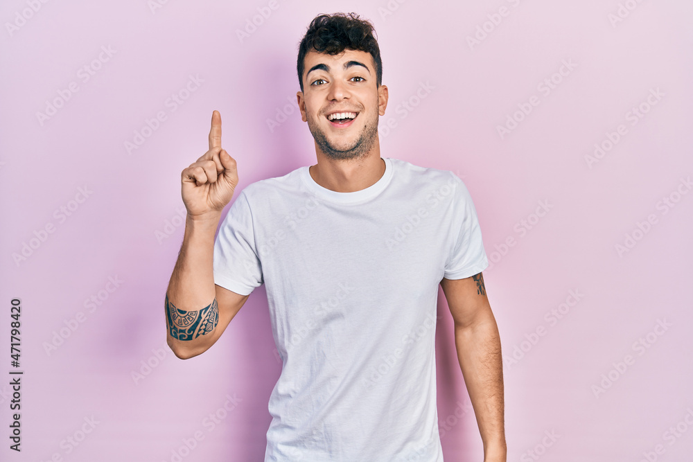 Young hispanic man wearing casual white t shirt smiling with an idea or question pointing finger up with happy face, number one Young hispanic man wearing casual white t shirt smiling with an idea or question pointing finger up with happy face, number one
