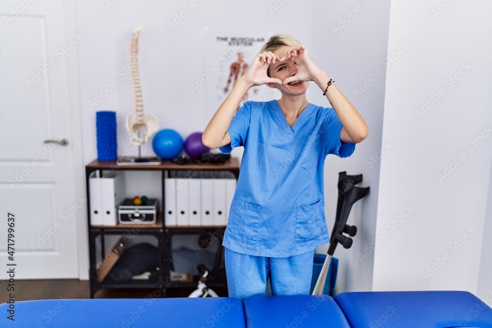 Young physiotherapist woman working at pain recovery clinic doing heart shape with hand and fingers smiling looking through sign Young physiotherapist woman working at pain recovery clinic doing heart shape with hand and fingers smiling looking through sign