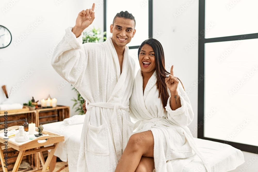 Obraz premium Young latin couple wearing towel standing at beauty center pointing finger up with successful idea. exited and happy. number one.