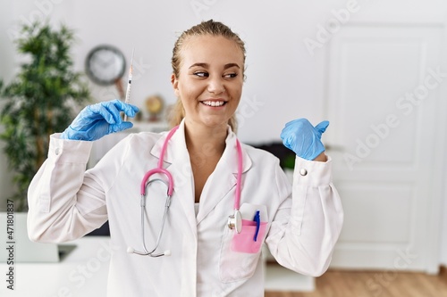 Young doctor woman holding syringe at the clinic pointing thumb up to the side smiling happy with open mouth