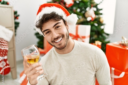 Young hispanic man smiling happy wearing christmas hat drinking champagne at home.