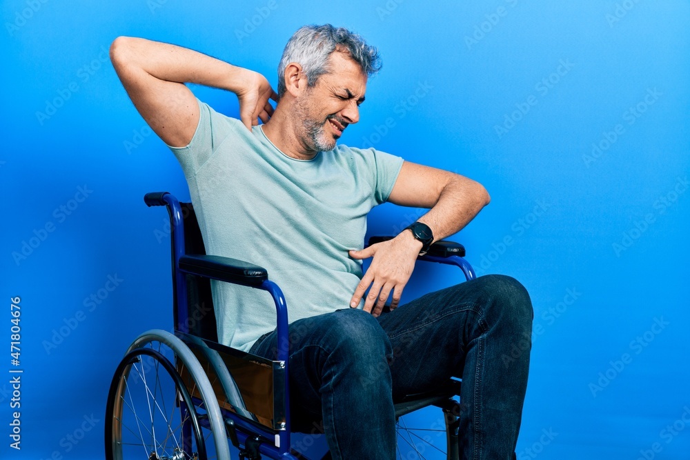Fototapeta premium Handsome middle age man with grey hair sitting on wheelchair suffering of neck ache injury, touching neck with hand, muscular pain