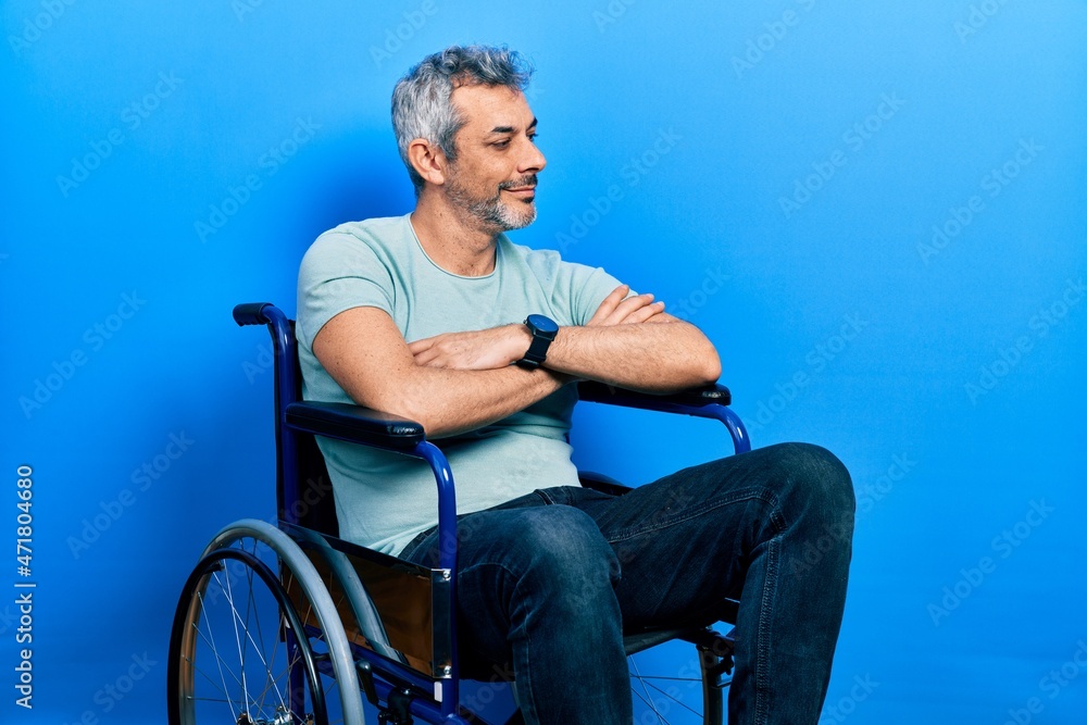 Fototapeta premium Handsome middle age man with grey hair sitting on wheelchair looking to the side with arms crossed convinced and confident