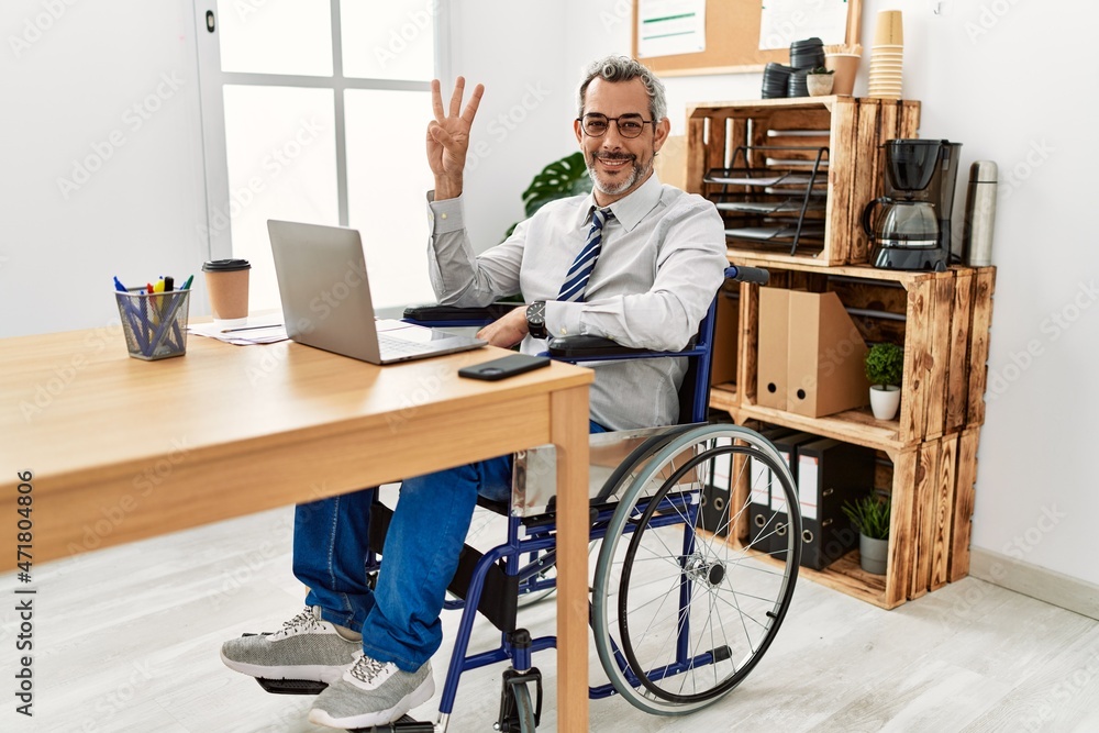 Middle age hispanic man working at the office sitting on wheelchair showing and pointing up with fingers number three while smiling confident and happy. Middle age hispanic man working at the office sitting on wheelchair showing and pointing up with fingers number three while smiling confident and happy.