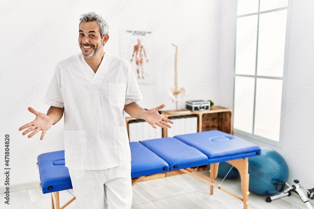 Fototapeta premium Middle age hispanic therapist man working at pain recovery clinic smiling cheerful with open arms as friendly welcome, positive and confident greetings