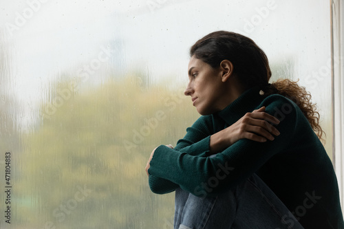 Raindrops like tears. Sad lonely latina female sit on windowsill in melancholic mood watch rain outside feeling her heart broken. Pensive young woman hug herself suffer of unanswered love. Copy space