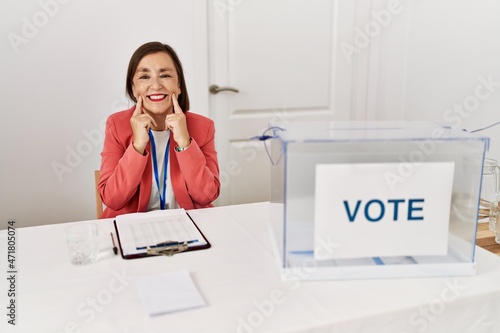 Beautiful middle age hispanic woman at political election sitting by ballot smiling with open mouth, fingers pointing and forcing cheerful smile photo