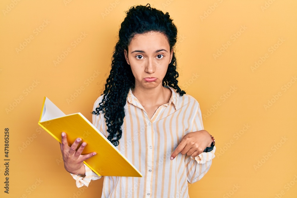 Young hispanic woman with curly hair holding book pointing down looking sad and upset, indicating direction with fingers, unhappy and depressed. Young hispanic woman with curly hair holding book pointing down looking sad and upset, indicating direction with fingers, unhappy and depressed.