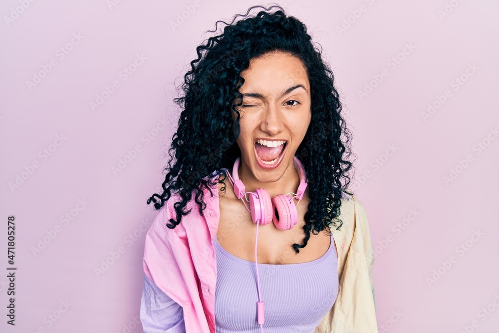 Young hispanic woman with curly hair wearing gym clothes and using headphones winking looking at the camera with sexy expression, cheerful and happy face. Young hispanic woman with curly hair wearing gym clothes and using headphones winking looking at the camera with sexy expression, cheerful and happy face.