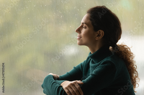 Music of rain. Side shot of serene latin woman relax by window with closed eyes listen sound of raindrops feel pleasure. Calm young lady enjoy breathing fresh ozonized air after rainstorm. Copy space.