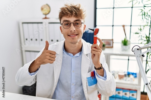 Young caucasian doctor man holding ear otoscope at the clinic smiling happy and positive, thumb up doing excellent and approval sign