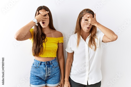 Mother and daughter together standing together over isolated background peeking in shock covering face and eyes with hand, looking through fingers with embarrassed expression.