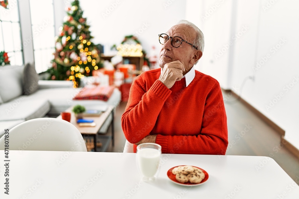 Senior man with grey hair sitting on the table with cookies by christmas tree serious face thinking about question with hand on chin, thoughtful about confusing idea Senior man with grey hair sitting on the table with cookies by christmas tree serious face thinking about question with hand on chin, thoughtful about confusing idea