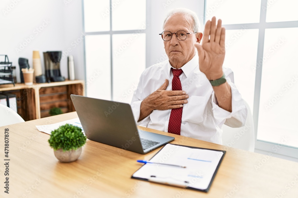 Senior man working at the office using computer laptop swearing with hand on chest and open palm, making a loyalty promise oath Senior man working at the office using computer laptop swearing with hand on chest and open palm, making a loyalty promise oath
