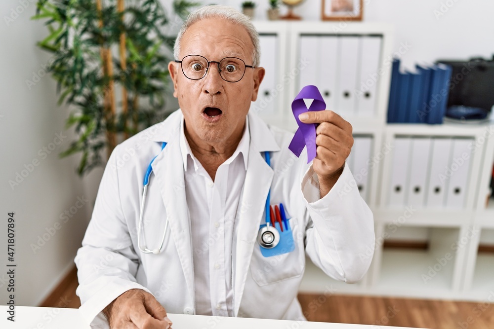 Senior doctor man holding purple ribbon awareness scared and amazed with open mouth for surprise, disbelief face Senior doctor man holding purple ribbon awareness scared and amazed with open mouth for surprise, disbelief face