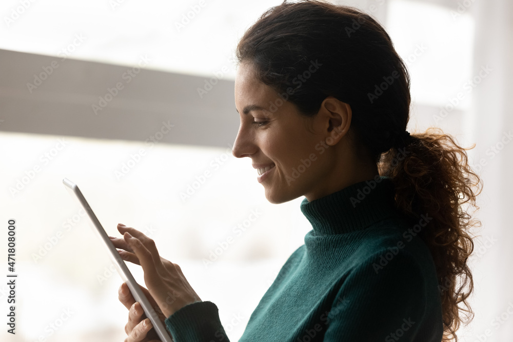 Digital pad user. Side view of smiling casual hispanic woman holding electronic tablet computer surfing social networks learning virtual course reading e book. Calm young female using tab pc by window - obrazy, fototapety, plakaty 