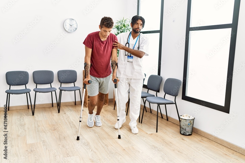 Two men physiptherapist and patient having medical consultation walking using crutches at hospital waiting room Two men physiptherapist and patient having medical consultation walking using crutches at hospital waiting room