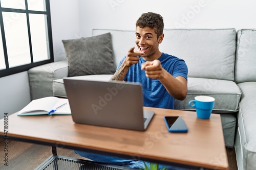Young handsome hispanic man using laptop sitting on the floor pointing fingers to camera with happy and funny face. good energy and vibes.