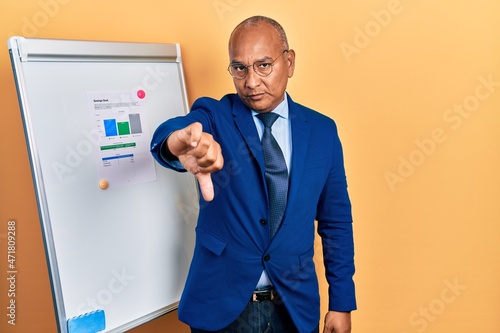 Middle age latin man wearing business clothes on chart presentation looking unhappy and angry showing rejection and negative with thumbs down gesture. bad expression.