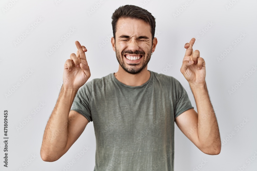 Young hispanic man with beard wearing casual t shirt over white background gesturing finger crossed smiling with hope and eyes closed. luck and superstitious concept. Young hispanic man with beard wearing casual t shirt over white background gesturing finger crossed smiling with hope and eyes closed. luck and superstitious concept.
