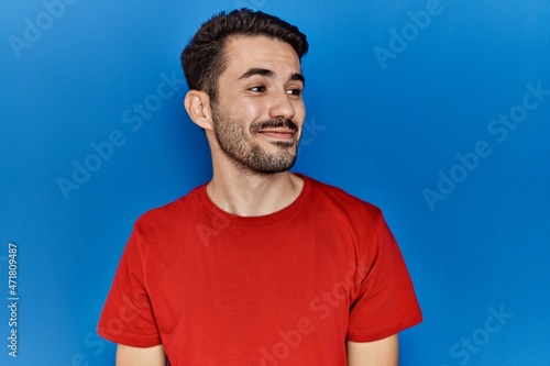 Young hispanic man with beard wearing red t shirt over blue background looking away to side with smile on face, natural expression. laughing confident.