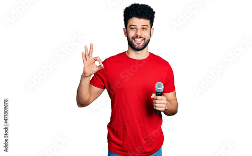 Young arab man with beard singing song using microphone doing ok sign with fingers, smiling friendly gesturing excellent symbol © Krakenimages.com