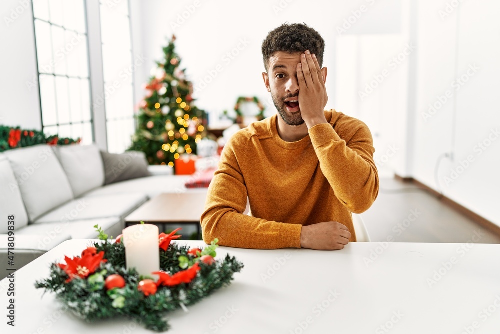 Arab young man sitting on the table by christmas tree covering one eye with hand, confident smile on face and surprise emotion. Arab young man sitting on the table by christmas tree covering one eye with hand, confident smile on face and surprise emotion.