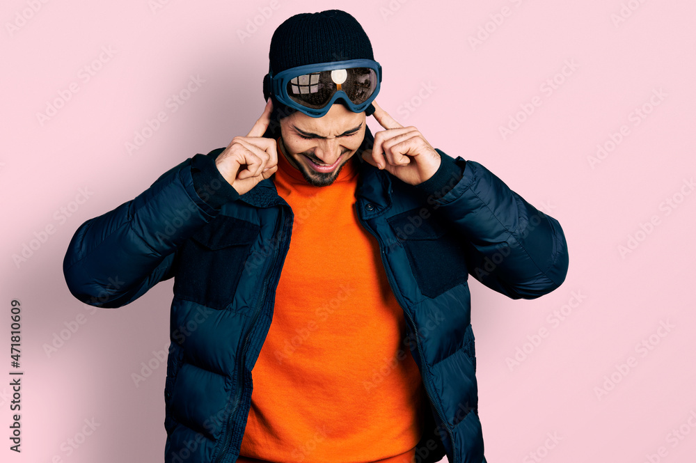 Young hispanic man with beard wearing snow wear and sky glasses covering ears with fingers with annoyed expression for the noise of loud music. deaf concept. Young hispanic man with beard wearing snow wear and sky glasses covering ears with fingers with annoyed expression for the noise of loud music. deaf concept.