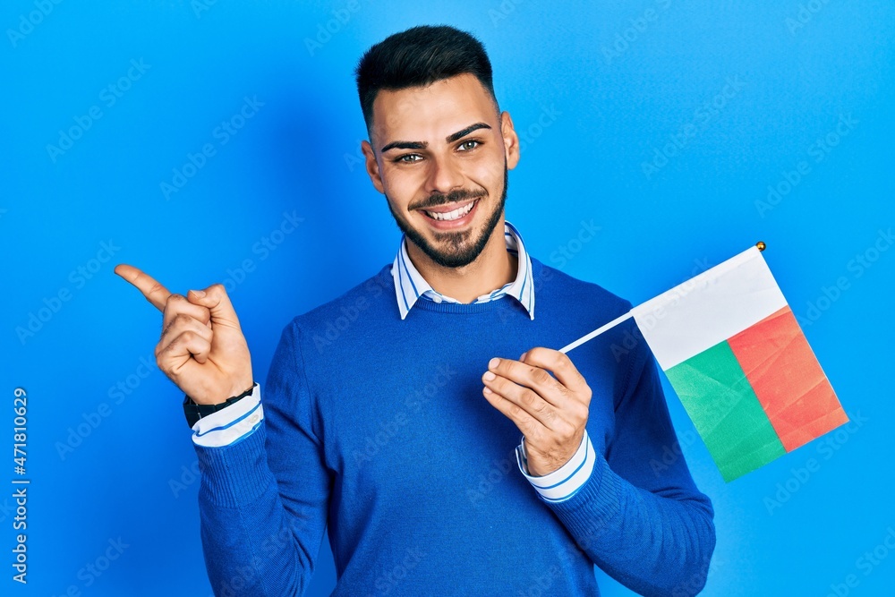 Young hispanic man with beard holding madagascar flag smiling happy pointing with hand and finger to the side
