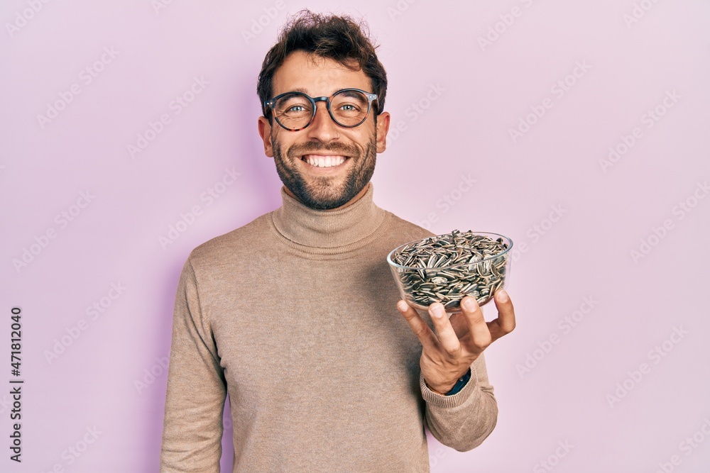 Handsome man with beard football reporter holding pumpkin seeds bowl looking positive and happy standing and smiling with a confident smile showing teeth - obrazy, fototapety, plakaty 
